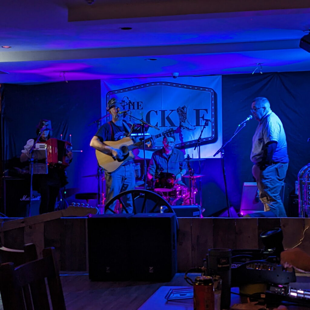 Picture of a gutarist, singer, accordion player, and a drummer performing  on stage at The Buckle